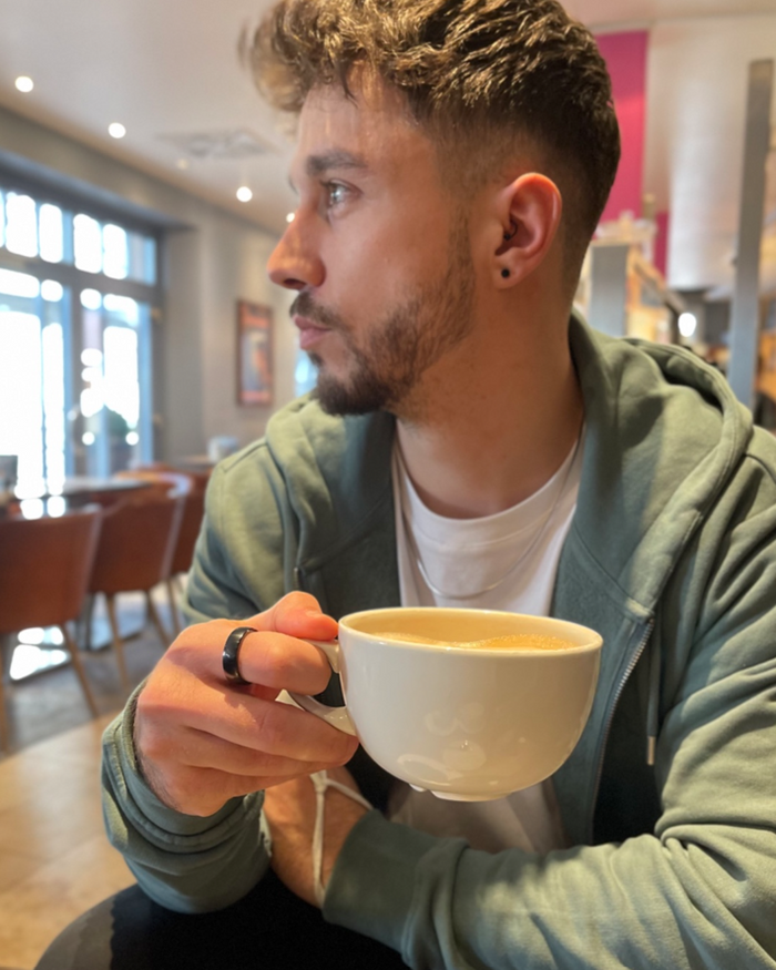 Uomo al bar con tazza di caffè in mano e PAGO Ring nero al dito anello di pagamento NFC sicuro per uso quotidiano